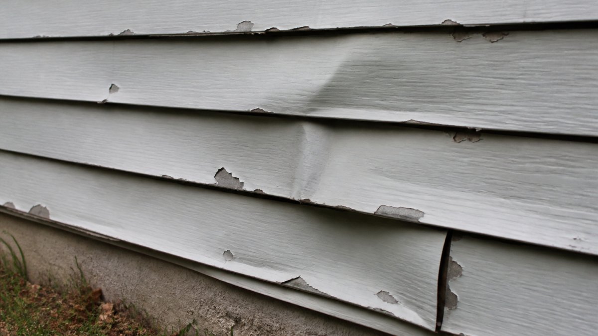 Close-up of damaged aluminum siding showing dents, warping, and peeling paint on a home's exterior wall in natural daylight